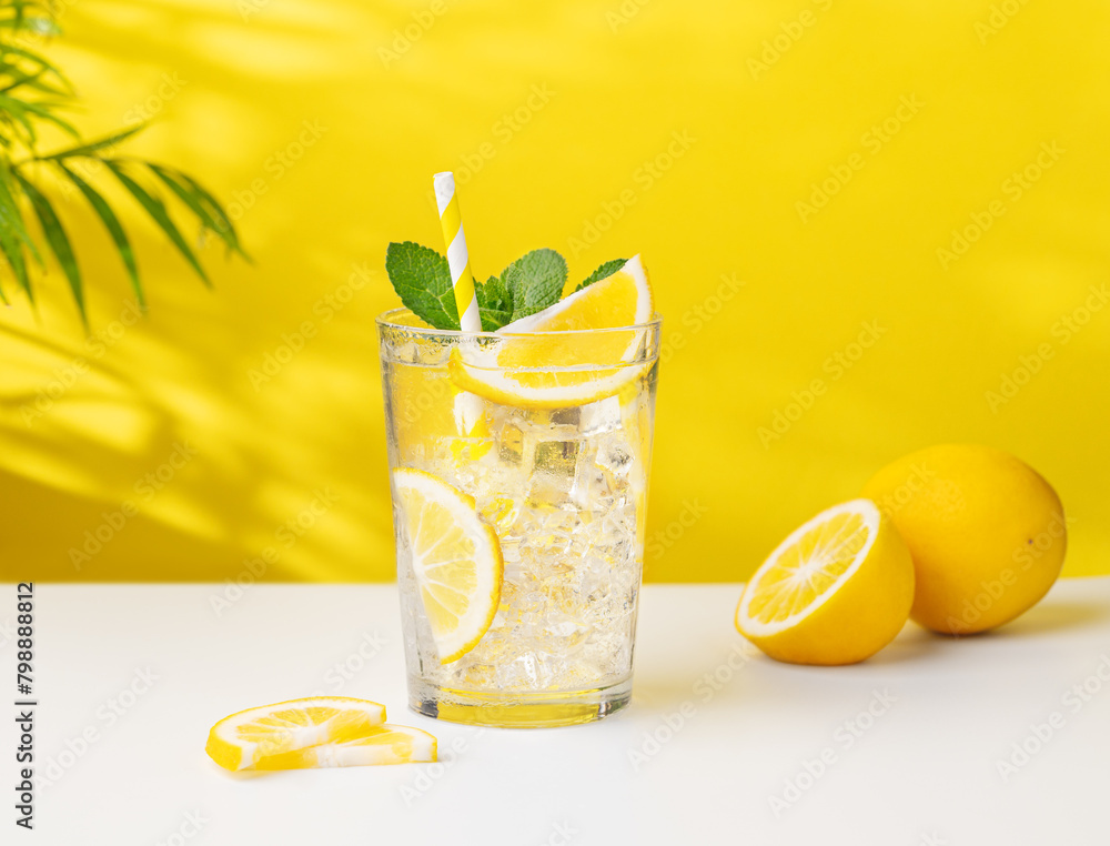 Φωτογραφία A glass of cold lemonade with lemon, mint and ice on a yellow and white background with a palm leaf and citrus fruits