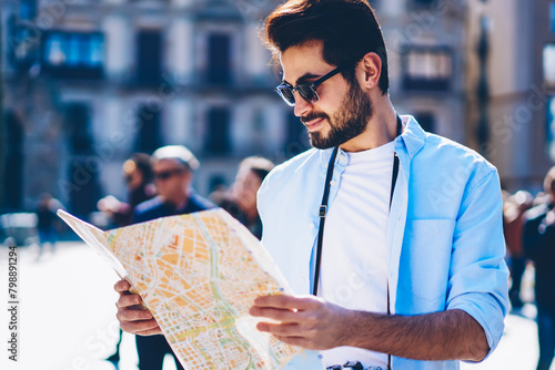 Bearded young tourist in sunglasses searching right direction on map enjoying wanderlust in new city.Casual dressed hister guy with vintage camera looking at route of showplaces standing outdoors photo