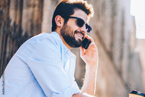 Cheerful bearded touris in sunglasses with cofffe to go in hand talking with friends in roaming on smartphone device sitting outdoors on street stairs.Positive man laughing during mobile conversation photo