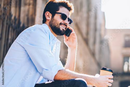 Cheerful bearded touris in sunglasses with cofffe to go in hand talking with friends in roaming on smartphone device sitting outdoors on street stairs.Positive man laughing during mobile conversation photo