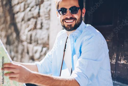 Cropped portrait of happy bearded tourist in stylish sunglasses holding travel map for searching showplaces in hands.Positive tourist in casual wear smiling at camera while sitting on street photo