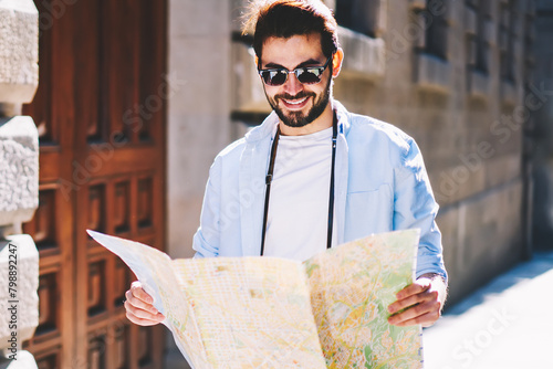 Positive bearded tourist happy to find right location of architectural showplaces on map strolling in uran setting in sunny day.Cheerful male traveler in stylish sunglasses walking on streets photo