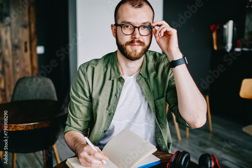 Young hipster male student in eyeglasses writing essay for homework bearded man thinking about tomorrow exam and looking aside, handsome creative author sitting at cafe interior with notebook photo