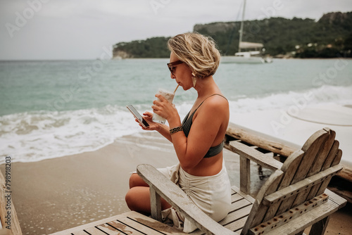 Young woman on the beach using mobile phone and drinking cocktail