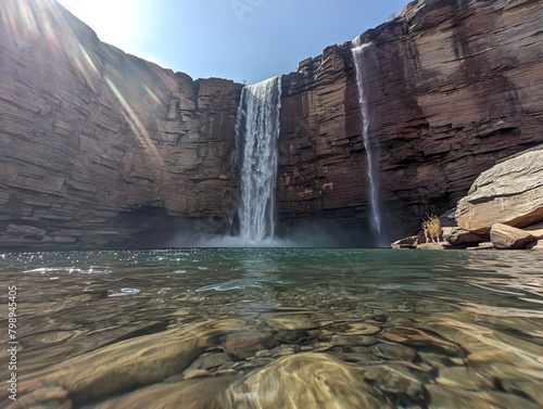 A waterfall is in the foreground of a large canyon. The water is clear and calm, and the rocks surrounding the waterfall are jagged and rough. The scene is peaceful and serene
