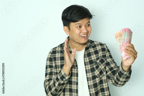 Young Asian man wearing brown flannel shirt showing shocked face expression while looking to paper money that he grab photo