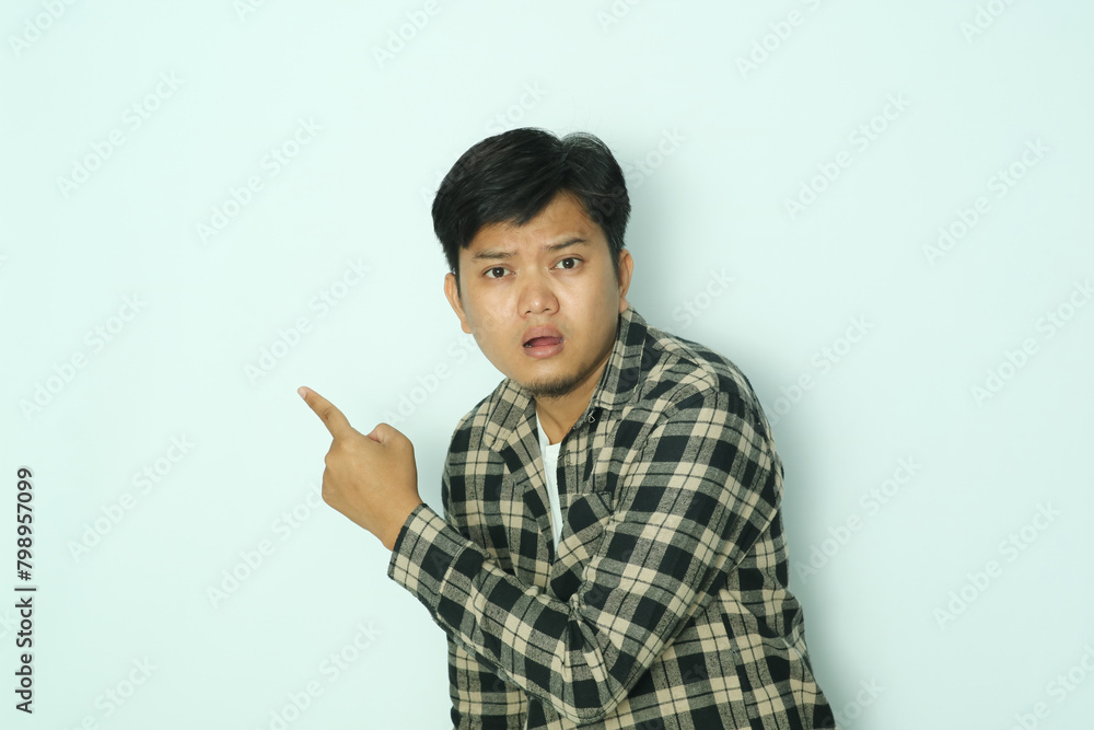 Young Asian man wearing brown flannel shirt looking camera with worried expression while pointing behind Young Asian man wearing brown flannel shirt looking camera with worried expression while pointing behind