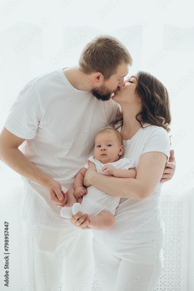 A happy young family mom dad and a small child smiling and kissing on a white isolated window background, a loving family threesome at home