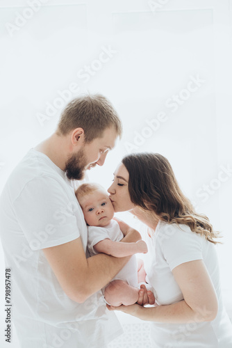 A happy young family mom dad and a small child smiling and kissing on a white isolated window background, a loving family threesome at home