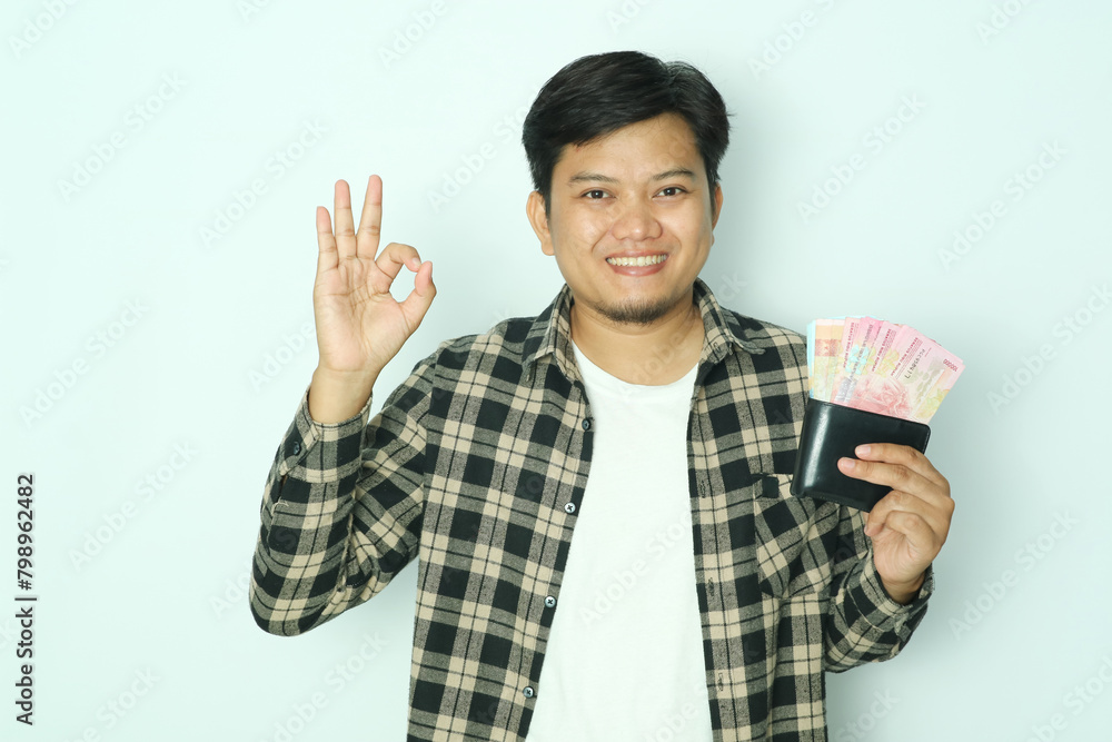 Young Asian man wearing brown flannel shirt smiling while holding wallet with money and give OK finger sign Young Asian man wearing brown flannel shirt smiling while holding wallet with money and give OK finger sign