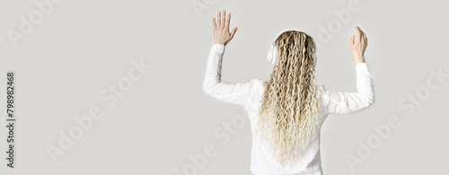 Modern stylish girl woman in headphones on head, listening to music and dancing, blonde long curly hair, back view, light gray background with copy space, minimal lifestyle no face trend photo