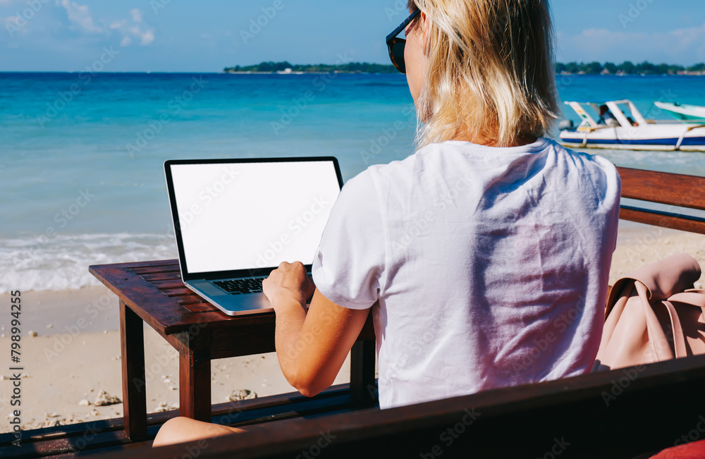 Back view of young blonde businesswoman working on ocean beach enjoying remote job in trip, hipster girl freelancer recreating near sea on summer typing publication on laptop computer for blog