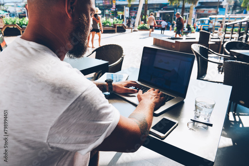 Back view of hipster guy sitting on street cafe with laptop computer typing feedback to friend in social networks, millennial male freelancer update content on website working remotely in cafeteria