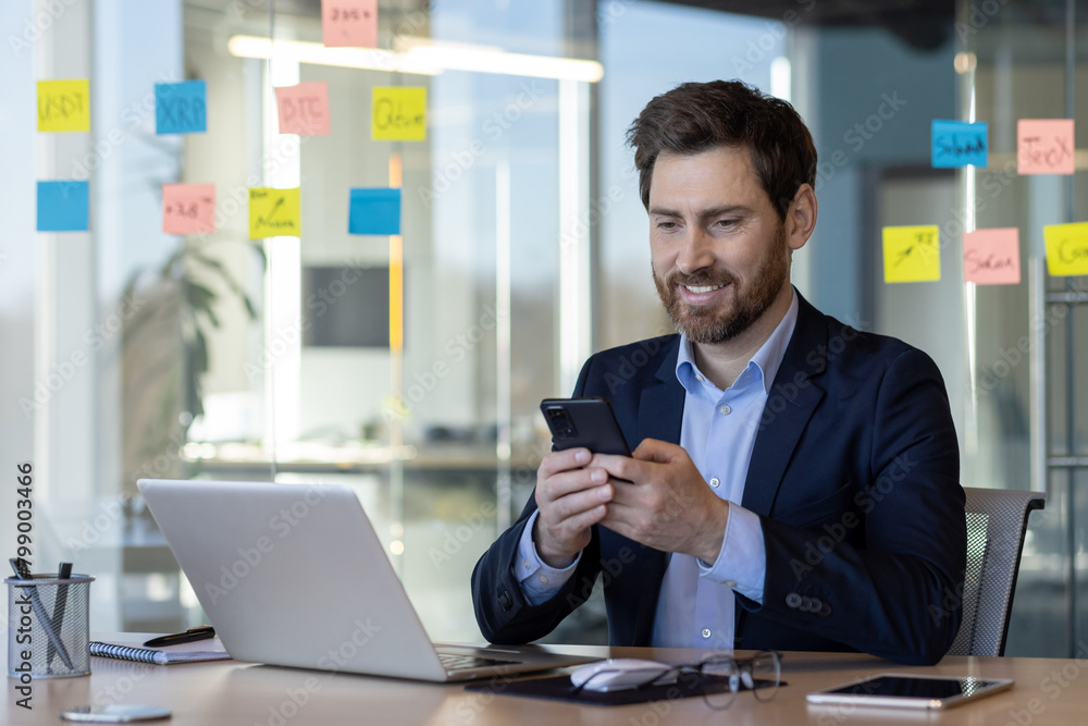 Satisfied smiling man using app on phone, businessman in business suit typing text message on smartphone, mature boss at workplace inside office with laptop, happy reading. Satisfied smiling man using app on phone, businessman in business suit typing text message on smartphone, mature boss at workplace inside office with laptop, happy reading.