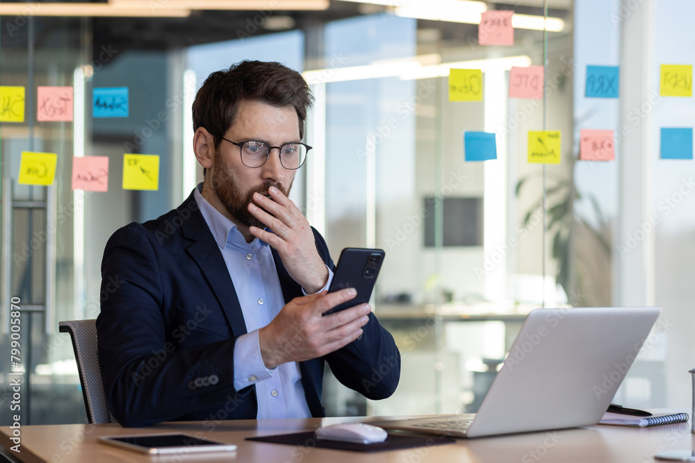 Sad disappointed businessman reading news on phone, man in business suit using app on smartphone, looking upset, received bad news online in email, working with laptop inside office. Sad disappointed businessman reading news on phone, man in business suit using app on smartphone, looking upset, received bad news online in email, working with laptop inside office.