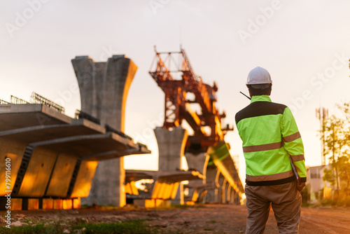 A construction worker is standing on a bridge, talking on his cell phone. The scene is set in a construction site, with a large crane in the background. The worker is wearing a yellow jacket © Chocheng channel