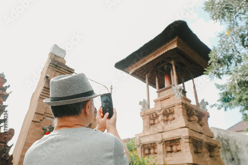 An adult man with grey hat is capturing photo of traditional Balinese Hindu temple with bokeh or blurred background