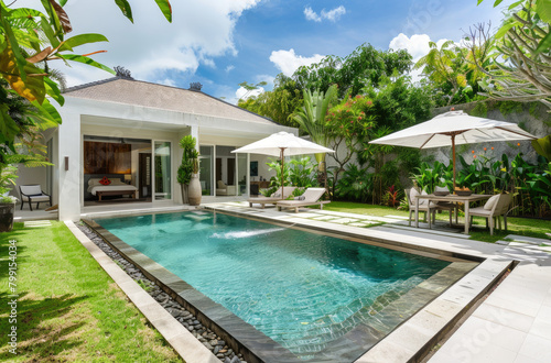 A modern and luxurious bungalow with an outdoor pool and lawn chairs under umbrellas, a dining table in front, surrounded by lush greenery and tropical plants