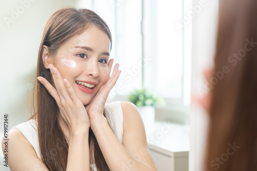 Facial beauty skin health care, smile of cute asian young woman looking at mirror, hand applying moisturizer lotion on her face, female putting cream treatment before makeup cosmetic routine at home.