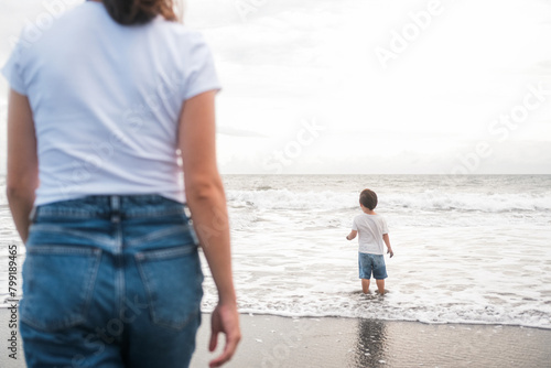 Mom and son Toddler are walking on the beach near the ocean. In the foreground is a silhouette of a woman in jeans watching her child splash in the water. Mothers Day.