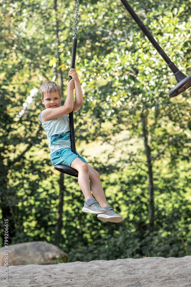 Vertical portrait of smiling little boy on swing in summer sunny forest