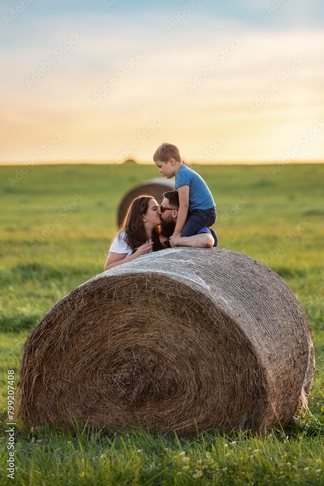 A happy family near a round roll of straw in a green meadow. The son is crouch high and watching his parents kiss A happy family near a round roll of straw in a green meadow. The son is crouch high and watching his parents kiss