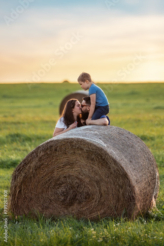 A happy family near a round roll of straw in a green meadow. The son is crouch high and watching his parents kiss
