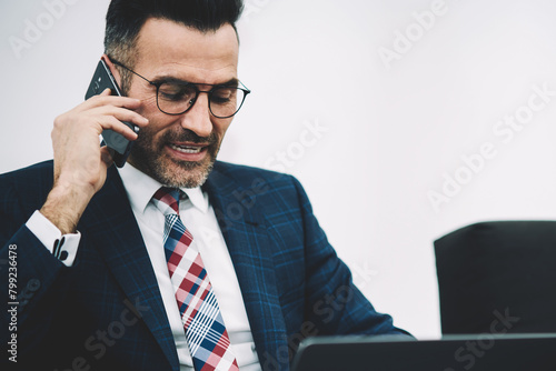 Serious mature entrepreneur in formal wear calling on smartphone talking with business partner working at modern netbook connected to wireless internet.Confident director communicating on cellular