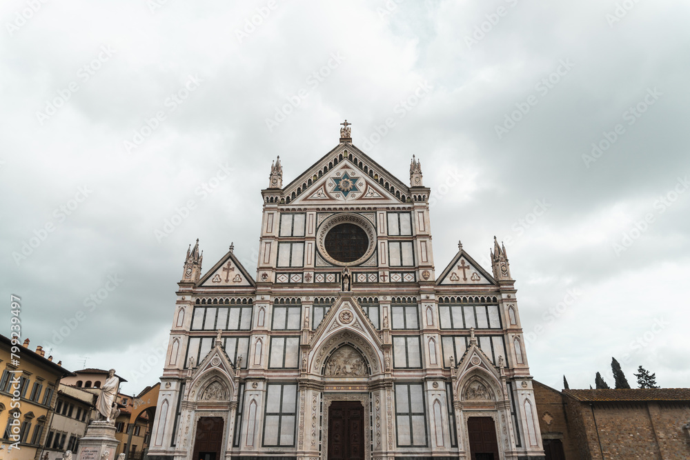 Basilica di Santa Croce's Neo-Gothic facade from Piazza di Santa Croce (Florence). Vibrant marble hues adorn three triangular pediments, echoing Florentine architectural tradition, under cloudy sky.