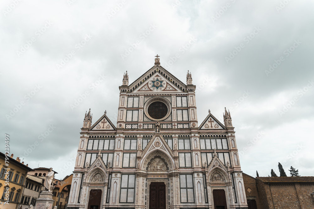 Basilica di Santa Croce's Neo-Gothic facade from Piazza di Santa Croce (Florence). Vibrant marble hues adorn three triangular pediments, echoing Florentine architectural tradition, under cloudy sky. Basilica di Santa Croce's Neo-Gothic facade from Piazza di Santa Croce (Florence). Vibrant marble hues adorn three triangular pediments, echoing Florentine architectural tradition, under cloudy sky.