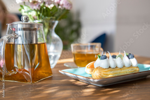 handmade eclairs on a plate, glass teapot with tea on a wooden table, Tea ceremony with cakes on the table and dark background.