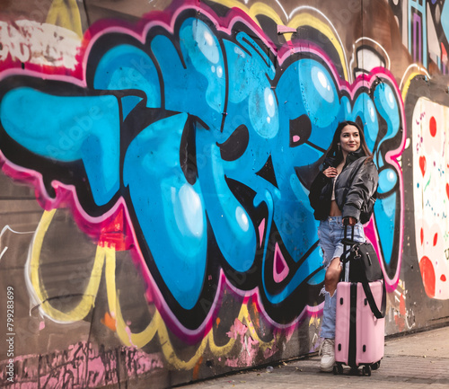 Urban stylish travel: young latina brunette with fashion look and pink suitcase in front of a lively street art mural at downtown