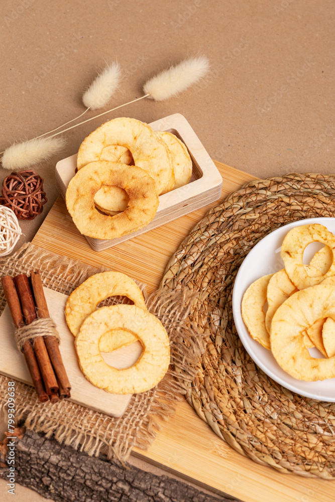 Fototapeta premium Crispy apple chips in a white round plate and in a wooden box, cinnamon sticks on a wooden desk on a beige background in rustic style. Healthy sweet snack.
