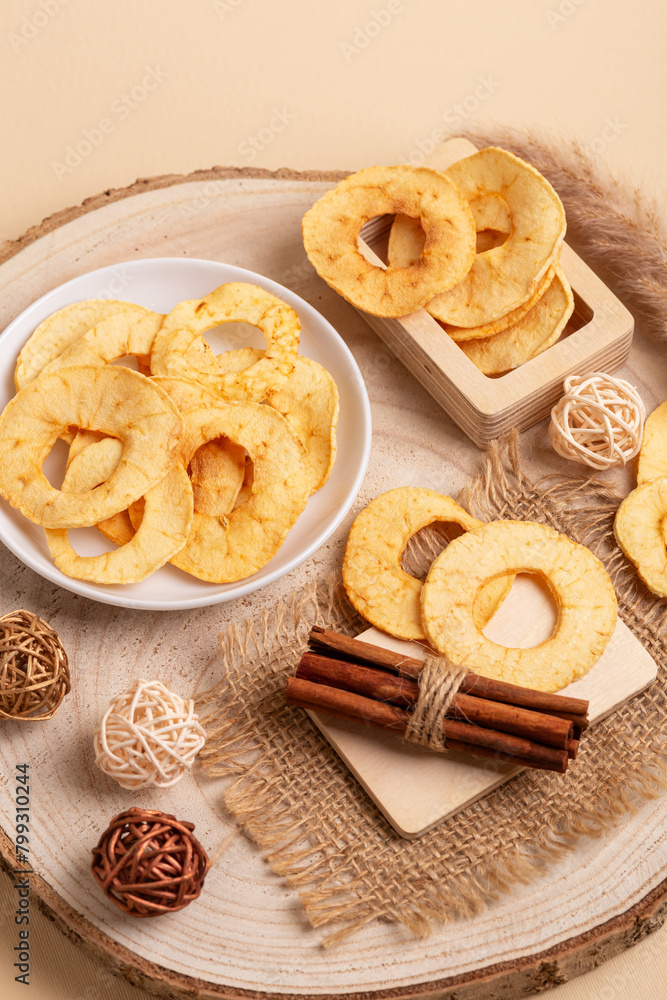 Tasty sweet apple chips in a plate and in a wooden box, cinnamon sticks on a wooden desk on a beige background. Tasty sweet apple chips in a plate and in a wooden box, cinnamon sticks on a wooden desk on a beige background.