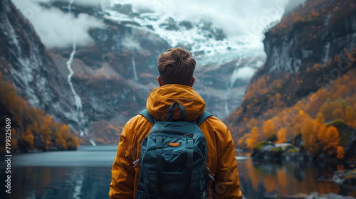 A man wearing a yellow jacket and backpack stands in front of a mountain range