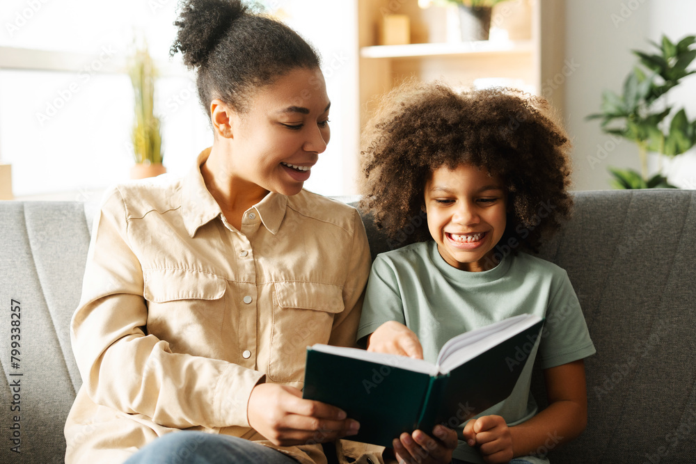 Happy young woman sitting next to her daughter on cozy sofa at home while reading book Happy young woman sitting next to her daughter on cozy sofa at home while reading book
