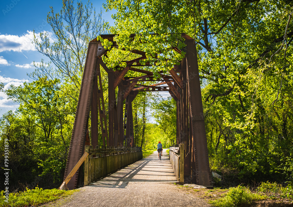 View of railroad bridge converted into recreational trail in summer; male bicyclist riding in background; lush greenery on both sides; blue sky View of railroad bridge converted into recreational trail in summer; male bicyclist riding in background; lush greenery on both sides; blue sky