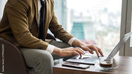 Businessman sitting on furniture working on laptop at ornamented corporate waiting area with cityscape background on the window. Business profession and strategic marketing plan for business success. © Summit Art Creations