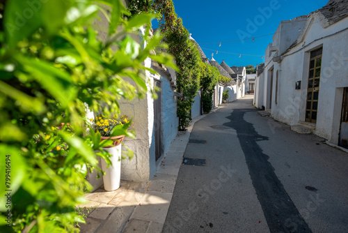 Beautiful stone of Trulli houses with white walls, narrow streets, decorated with flowers, palm trees and other decorations. Spring day, warm with sun and blue skies