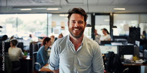 A smiling man with curly hair is standing confidently in the foreground of an office environment. He's wearing a casual collared shirt and appears to be a business professional or small business owner