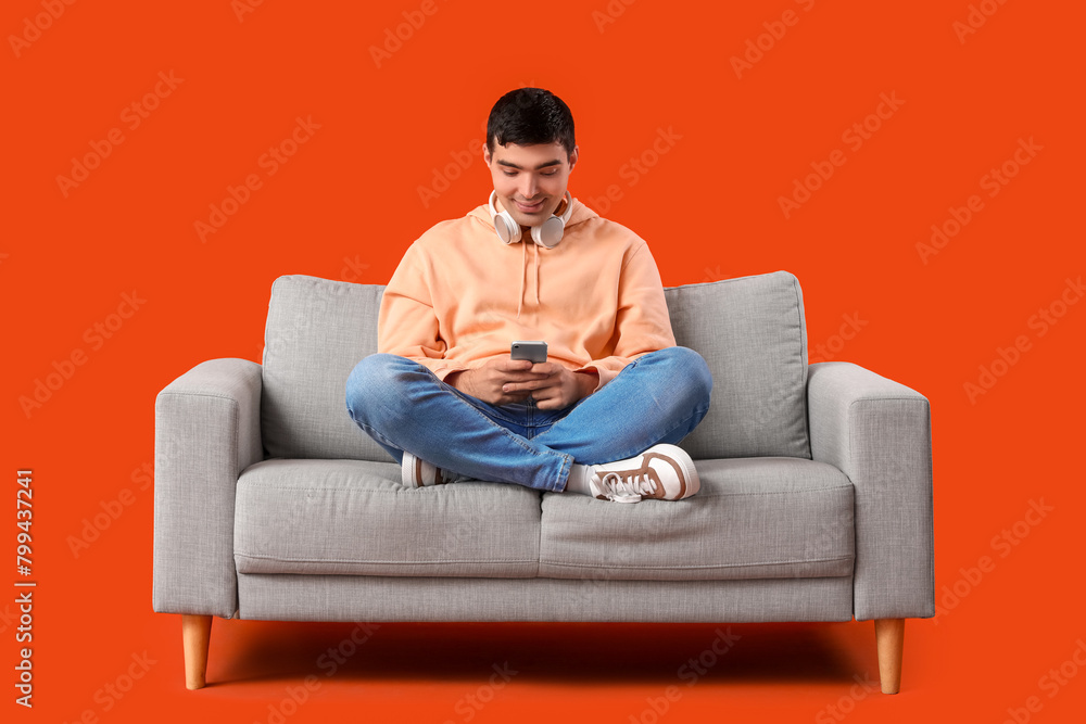 Handsome young man using mobile phone on grey sofa against orange background Handsome young man using mobile phone on grey sofa against orange background