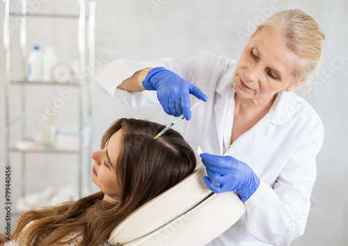 Old woman doctor implementing hair care procedure by injection method for patient lying on clinical chair