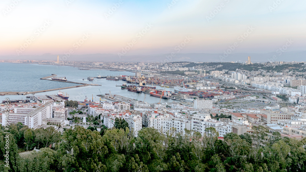 Algiers sea bay and pier port aerial view on the capital city buildings, trees and Mediterranean blue water ships and boats. Colorful sky by sunset. The Great mosque and the Martyr's Memorial monument