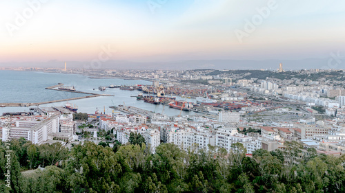 Algiers sea bay and pier port aerial view on the capital city buildings, trees and Mediterranean blue water ships and boats. Colorful sky by sunset. The Great mosque and the Martyr's Memorial monument