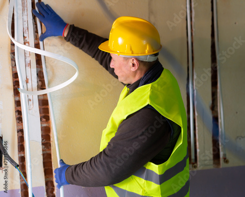 Caucasian electrician in vest and hardhat lying down cable duct during repair works in apartment. photo