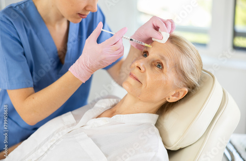 Old female patient lying on clinical chair undergoing face care procedure by injection method