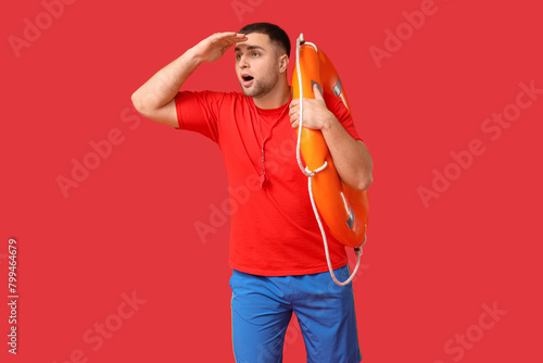 Worried young lifeguard with whistle and lifebuoy looking at somewhere on red background