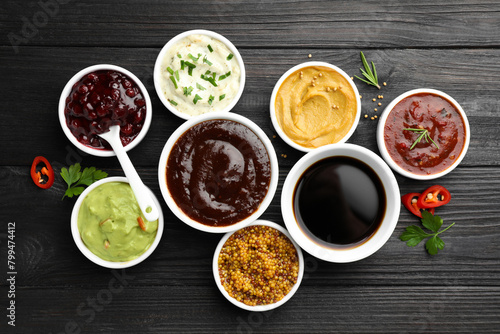Different tasty sauces in bowls, parsley, chili pepper and rosemary on black wooden table, flat lay