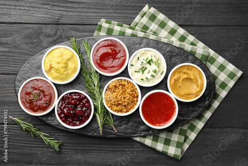 Different tasty sauces in bowls and rosemary on black wooden table, top view