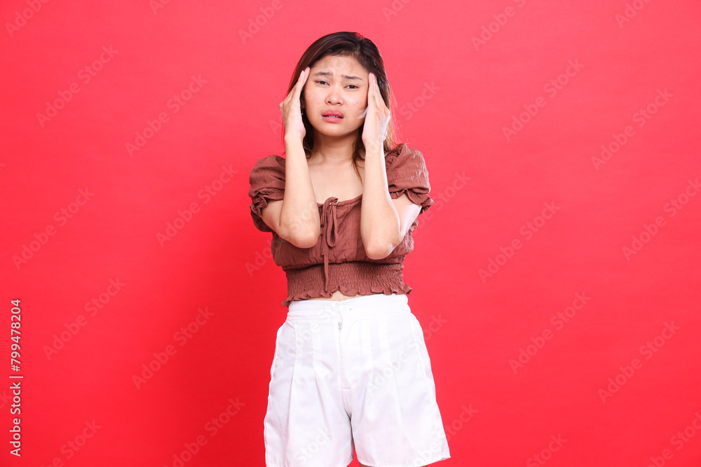 Asian woman's expression with both hands holding her head forward, very painful, dizzy, candid, wearing a brown blouse and shorts with a red background. for health, care and advertising concept Asian woman's expression with both hands holding her head forward, very painful, dizzy, candid, wearing a brown blouse and shorts with a red background. for health, care and advertising concept